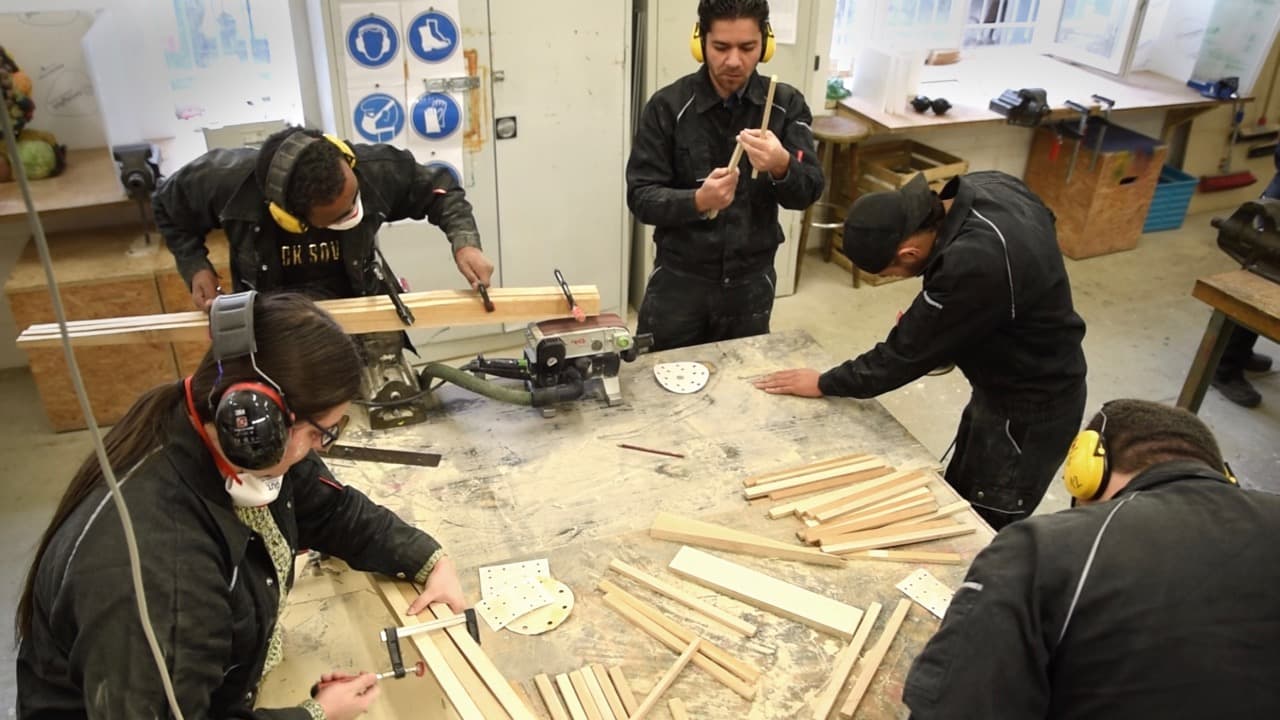 Das Bild zeigt fünf Männer. Sie befinden sich in einer Schreinerei und tragen alle Kopfhörer. Sie arbeiten an einer Werkbank an kleinen Holzscheiten. 