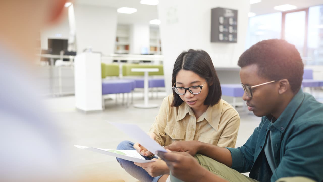 Das Bild zeigt eine Studentin und einen Studenten. Sie sitzen zusammen und schauen sich Formulare an. Im hintergrund sieht man eine Sitzugruppe mit Tischen und Bänken.