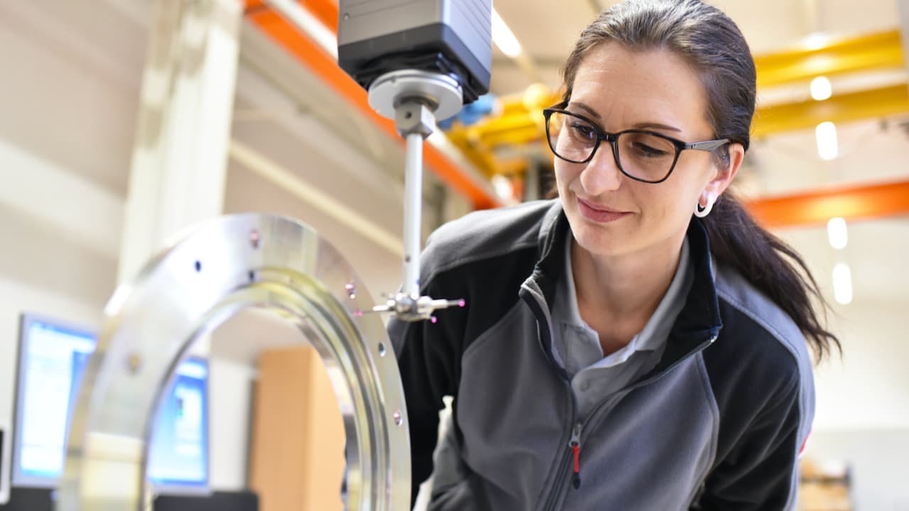 Das Bild zeigt eine Frau, die sich zu einer Maschine hinunterbeugt. Im Hintergrund sieht man einen Computer und Rohre.