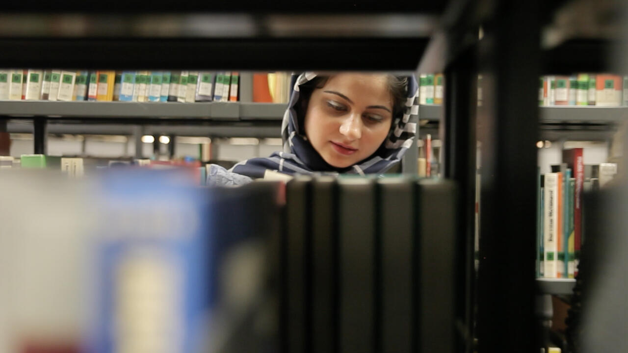 Das Bild zeigt eine junge Frau mit Kopftuch in einer Bibliothek. Ihr Kopf spitzt hinter einem Bücherregal hervor.