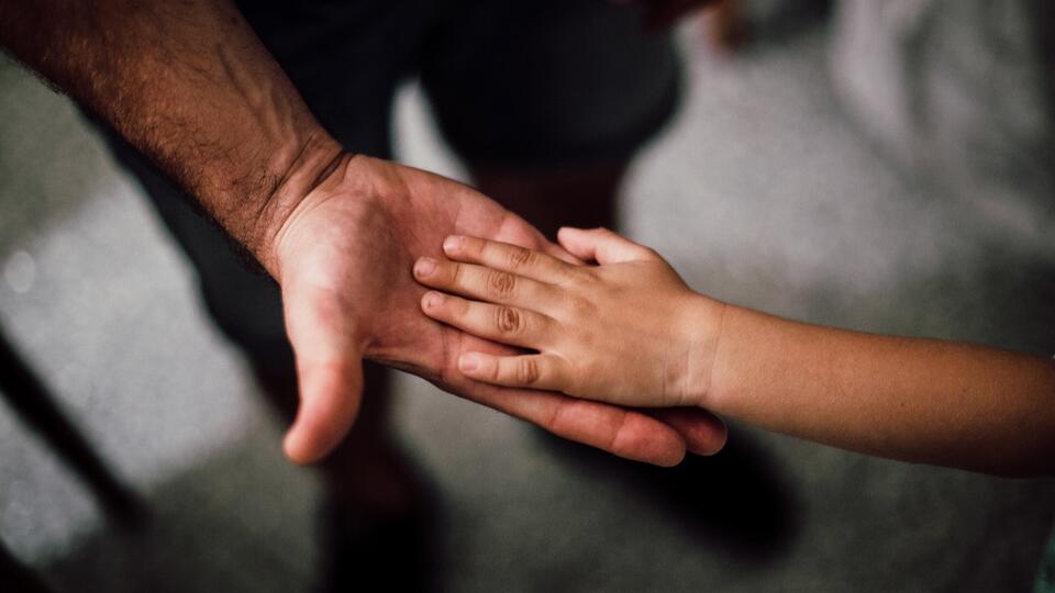 Das Bild zeigt die Hand eines Mannes und eines Kindes. Das Kind legt seine Hand in die Hand des Mannes.