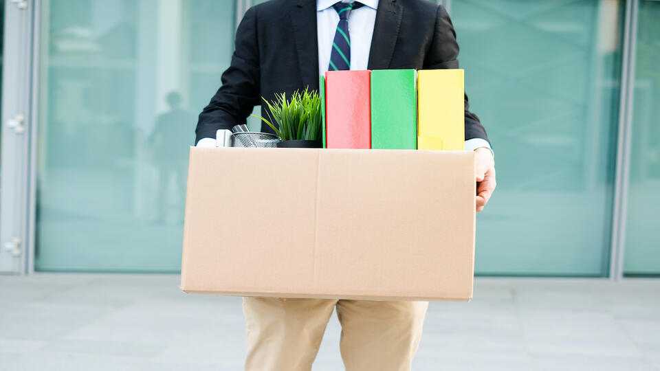 Das Bild zeigt einen Mann vor einem Bürogebäude. Er trägt einen Karton. Im Karton ist eine Pflanze und drei bunte Ordner.