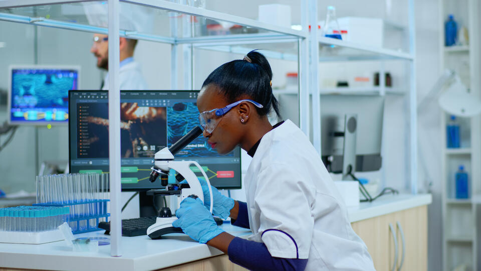 Das Bild zeigt eine Forscherin. Sie sitzt an einem Labortisch und untersucht eine Probe unter einem Vergrößerungsglas. Im Hintergrund sieht man einen PC. 