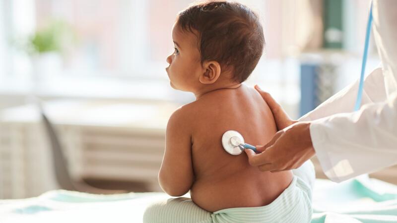 Das Bild zeigt ein Kleinkind mit nacktem Oberkörper. Es sitzt auf einer Untersuchungsliege. Eine Ärztin untersucht es mit einem Stethoskop. 