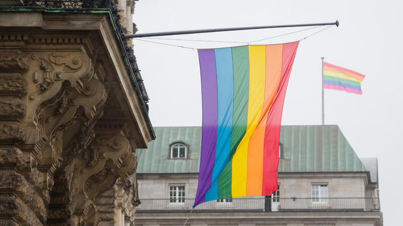 Das Bild zeigt die Regenbogenflagge. Sie hängt von einem älteren Gebäude herab.