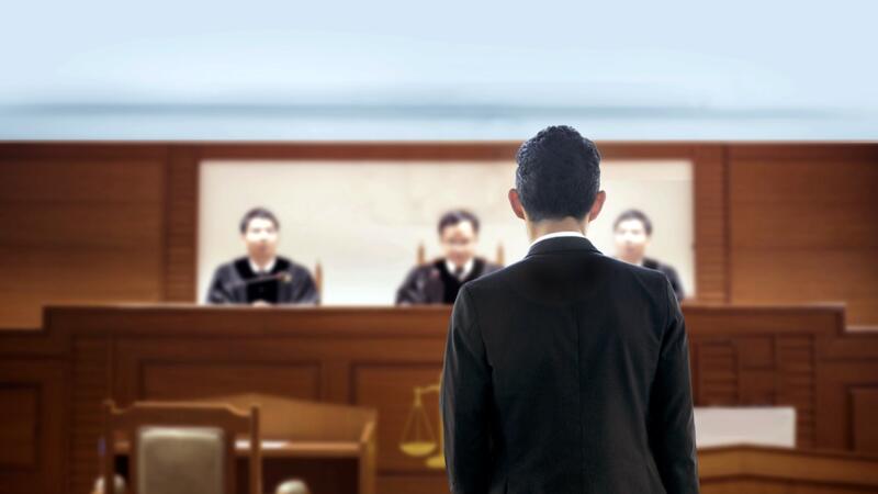 Das Bild zeigt einen Mann von hinten. Er steht vor dem Richterpult. Hinter dem Richterpult sitzen drei Richter.