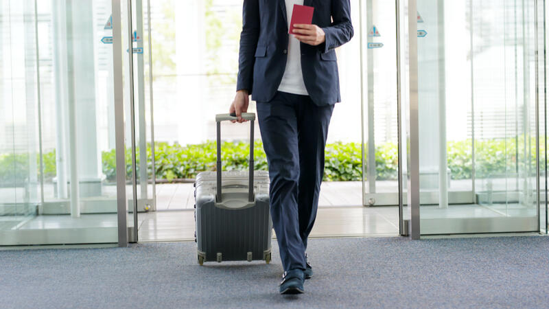 Das Bild zeigt einen Mann im Anzug. Er betritt gerade die Lobby eines Gebäudes und zieht einen grauen Rollkoffer hinter sich her. in der Hand hält er einen roten Reisepass.