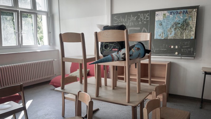 Klassenraum mit gestapelten Tischen und Stühlen, Schultüte und Rucksack vor einer Tafel.